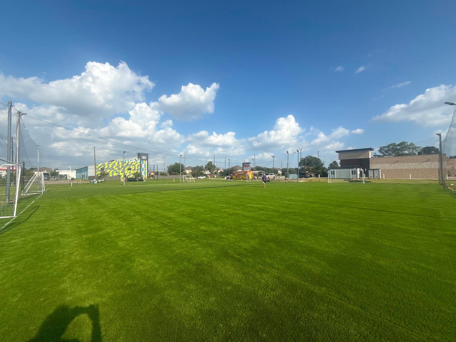 The same 7-a-side turf pitches at Turf On Soccer Fields in Houston, TX in daylight — green artificial turf, goals at both ends and perimeter netting under a blue sky.