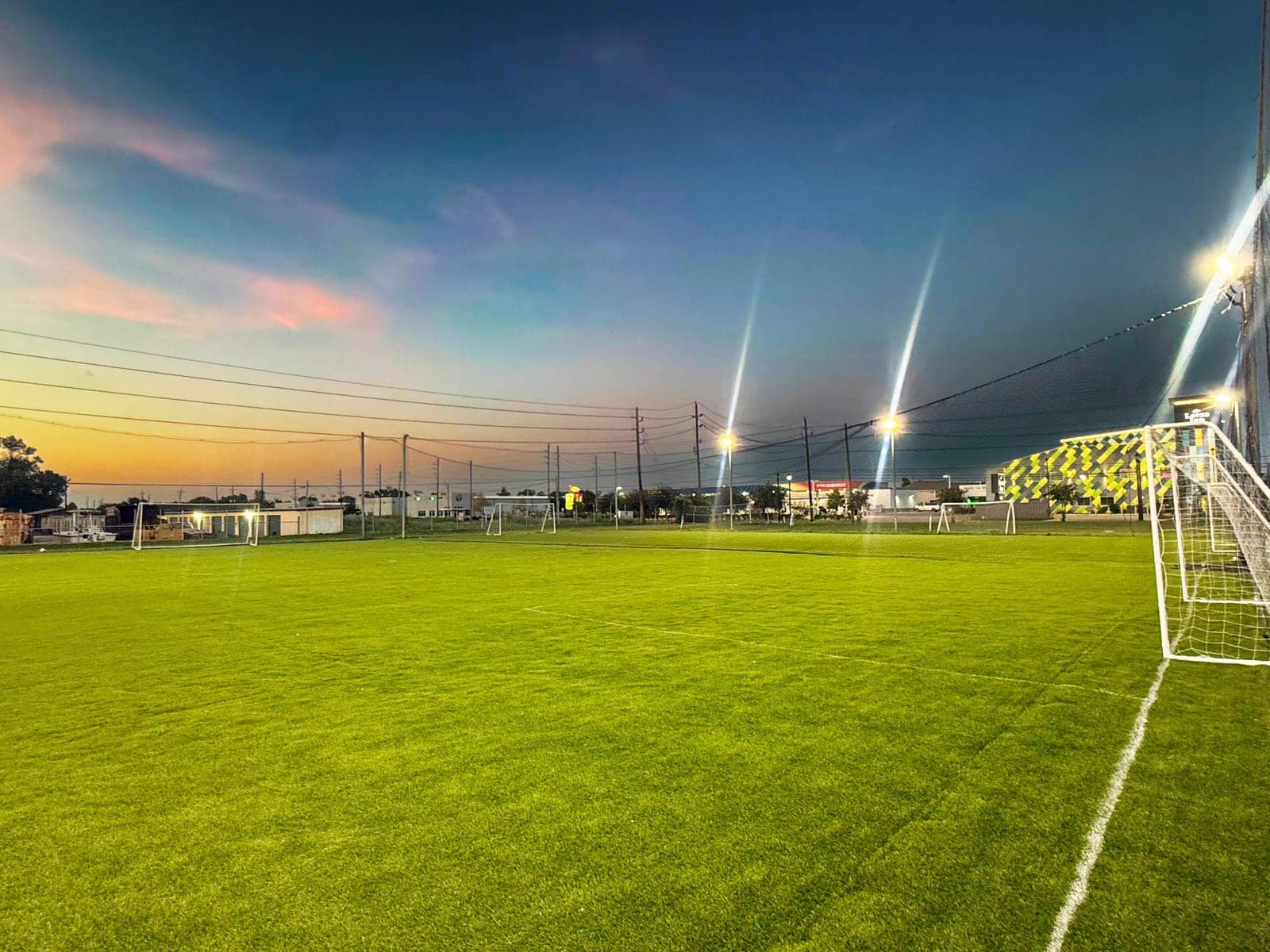 The 7-a-side turf pitches at Turf On Soccer Fields in Houston, TX at dusk, floodlights lit over green artificial turf, goals and white line markings under an orange sunset sky.